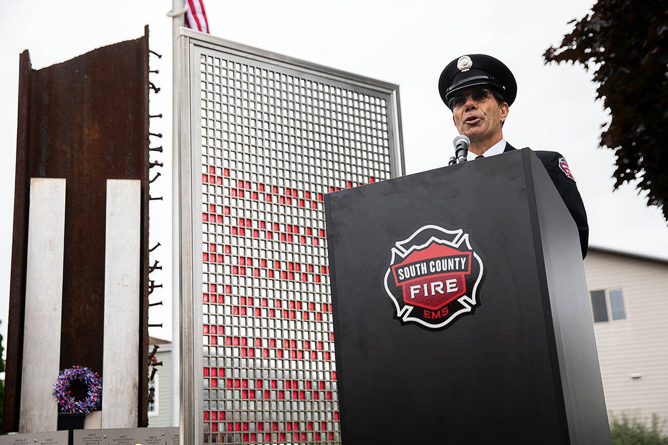 Retired South County Firefighter Dave Erickson speaks to a crowd of 50 people gathered outside of the Fallen Firefighter Memorial Park at the downtown Edmonds Fire Station on Wednesday, Sept. 11, 2024 for a 9/11 Memorial Ceremony. In the background of the ceremony stands a 1-ton beam recovered from the collapsed World Trade Center along with multicolored glass tiles. The tiles represent the more than 3,000 people killed, including 343 firefighters, 60 police and 10 emergency medical services workers. (Olivia Vanni / The Herald)